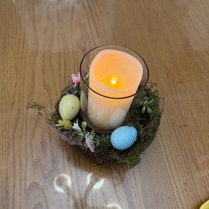 May include: A lit candle in a glass jar sits in a nest of twigs and moss with three pastel colored Easter eggs.