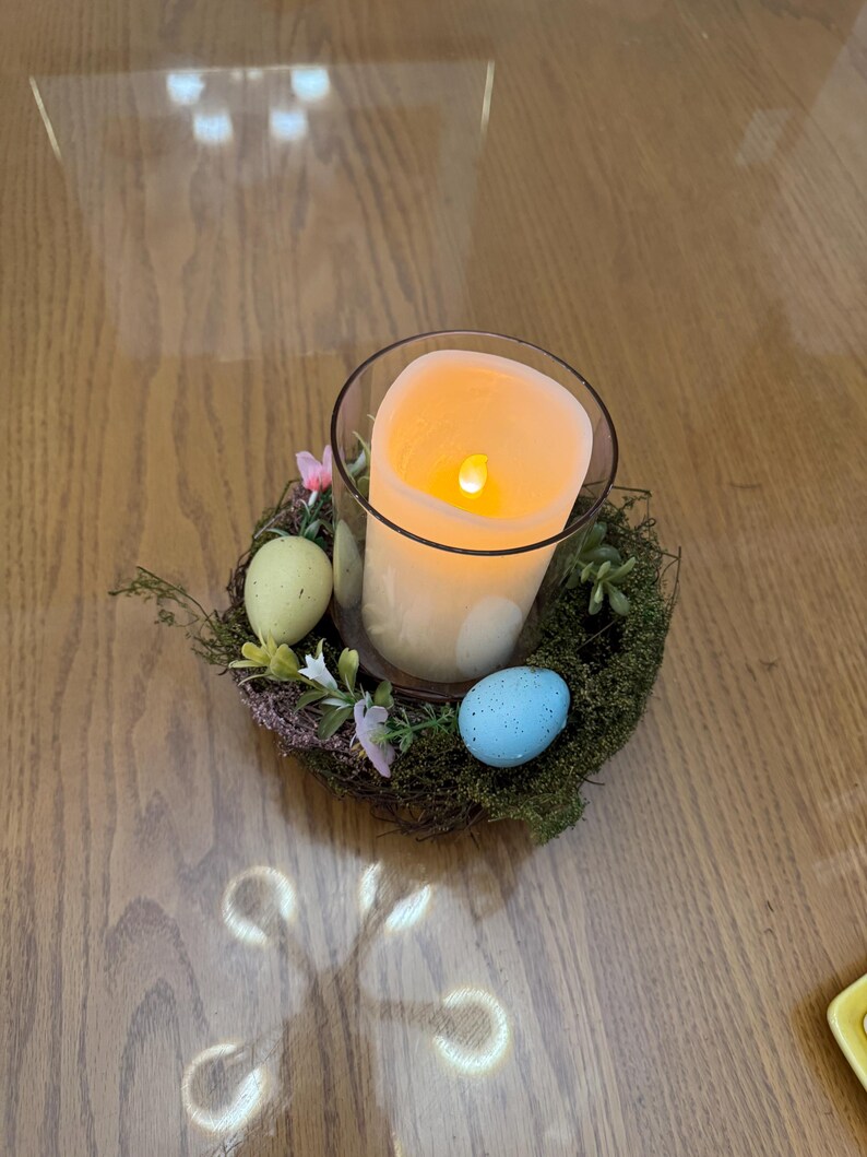 May include: A lit candle in a glass jar sits in a nest of twigs and moss with three pastel colored Easter eggs.
