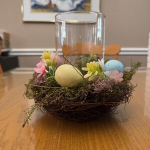 May include: A brown woven bird's nest with green moss and pink and white flowers. Three pastel colored Easter eggs are nestled in the nest.