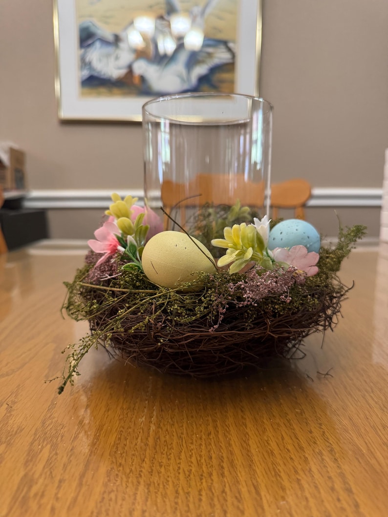May include: A brown woven bird's nest with green moss and pink and white flowers. Three pastel colored Easter eggs are nestled in the nest.