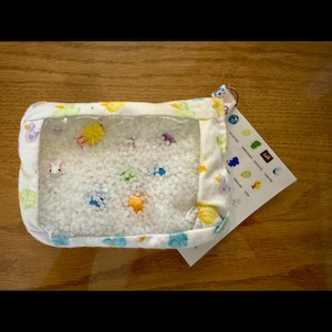 May include: A white fabric sensory toy with a clear plastic window. The window shows white beads and colourful small toys, including a yellow chick, a blue butterfly, a green frog, and a white bunny. The fabric has a colourful pattern of chicks, butterflies, frogs, and bunnies.