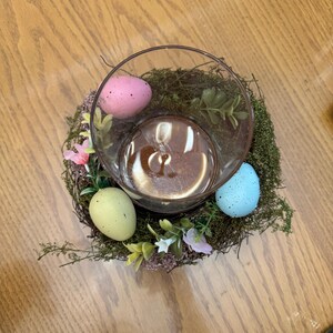 May include: A glass vase with a nest of moss and artificial flowers, decorated with pastel colored speckled Easter eggs.