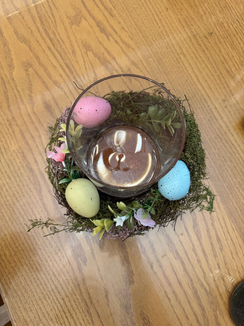 May include: A glass vase with a nest of moss and artificial flowers, decorated with pastel colored speckled Easter eggs.