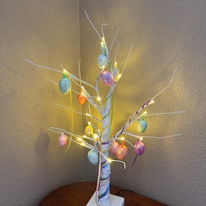 May include: A white, twig-like tree with colorful Easter eggs hanging from the branches. The tree is decorated with white lights and has a white base.