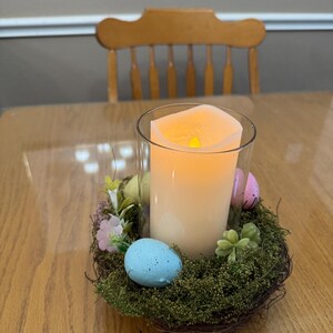 May include: A lit candle in a glass jar sits in a nest of twigs and moss. The nest is decorated with pastel colored Easter eggs and small green plants.