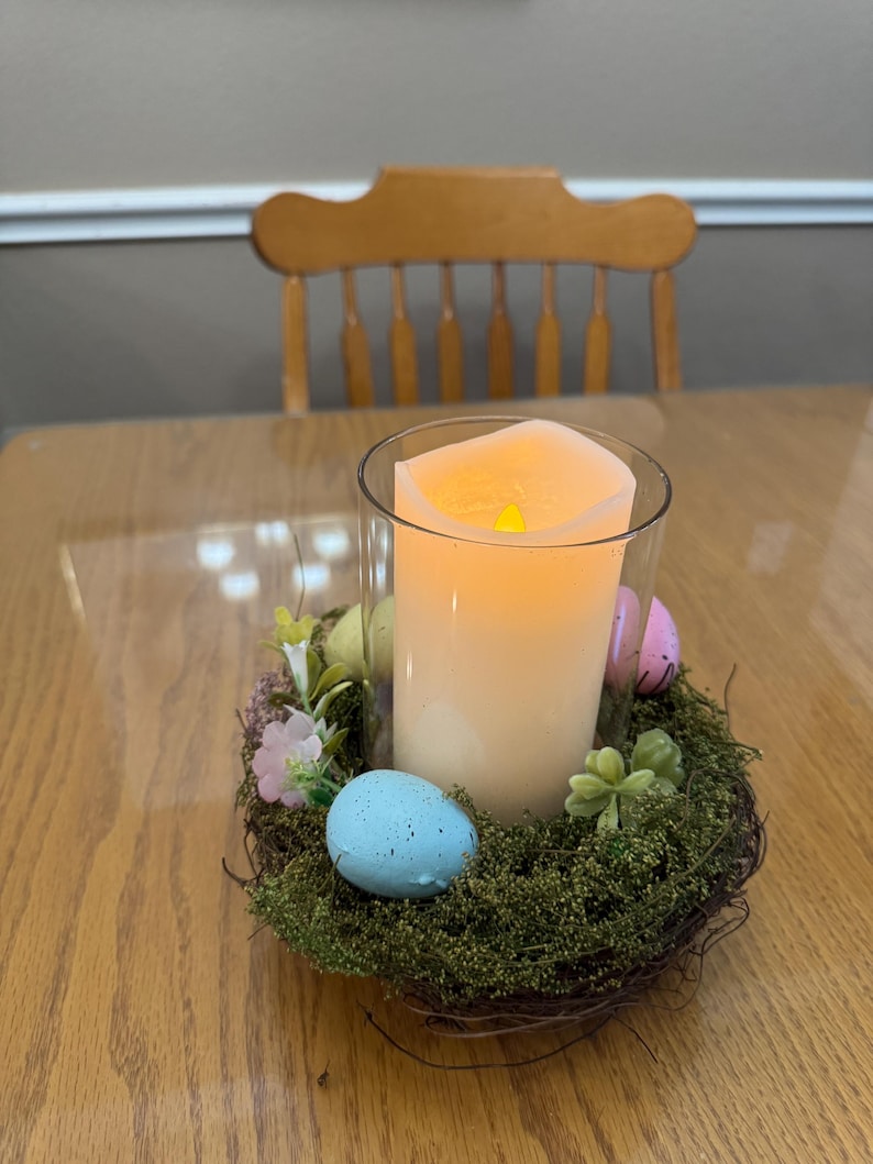 May include: A lit candle in a glass jar sits in a nest of twigs and moss. The nest is decorated with pastel colored Easter eggs and small green plants.