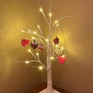 May include: A decorative white birch tree with warm white LED lights and heart-shaped ornaments. The tree is on a white square base and sits on a wooden surface. The ornaments are red and clear.