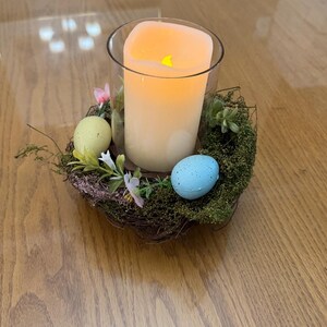May include: A glass candle holder with a white pillar candle inside, surrounded by a nest of twigs and moss. There are three decorative Easter eggs, one blue and two yellow, nestled in the moss.