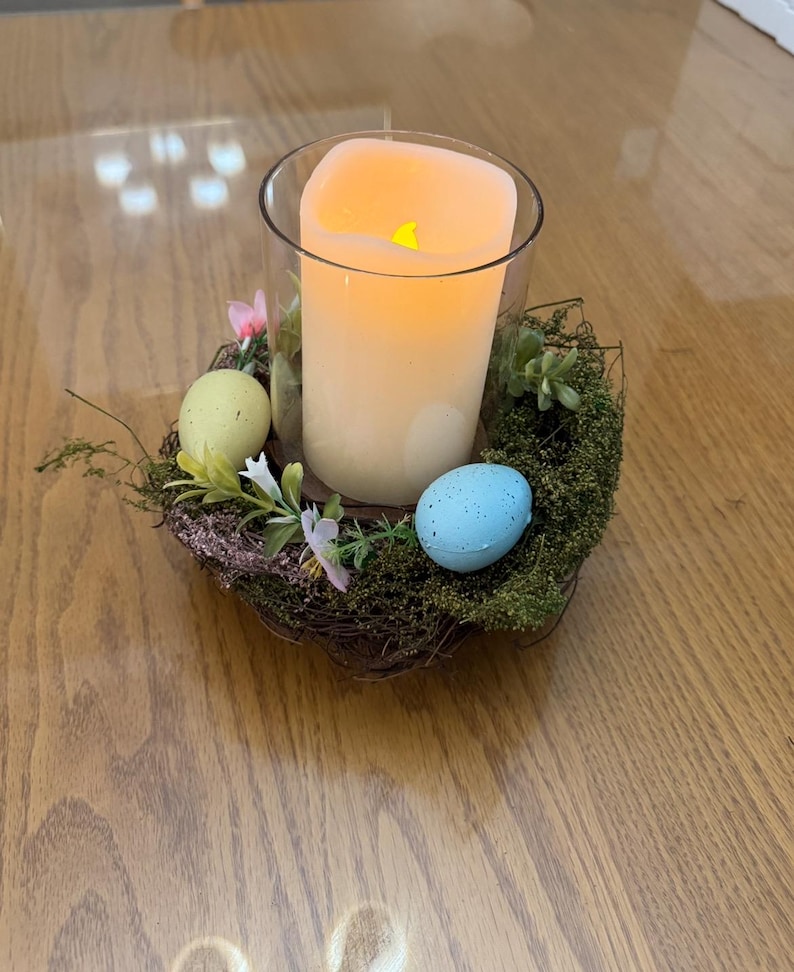 May include: A glass candle holder with a white pillar candle inside, surrounded by a nest of twigs and moss. There are three decorative Easter eggs, one blue and two yellow, nestled in the moss.
