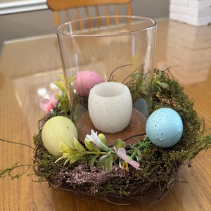 May include: A glass candle holder with a white candle inside, surrounded by a nest of twigs and moss. There are four pastel colored Easter eggs around the candle holder.