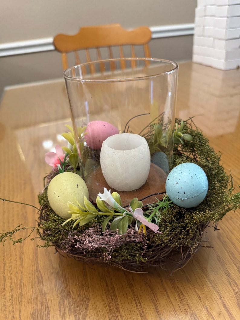 May include: A glass candle holder with a white candle inside, surrounded by a nest of twigs and moss. There are four pastel colored Easter eggs around the candle holder.