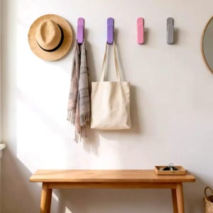 May include: A minimalist wall arrangement with four coloured hooks. A straw hat, a patterned scarf, and a canvas tote bag are suspended from the hooks. A wooden bench and a circular mirror add to the simple decor. Hooks are purple, pink and grey.