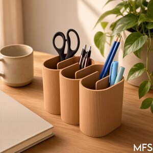 May include: A light tan desk organiser with a vertical ribbed texture and three interconnected oval compartments. The organiser holds black scissors, two pens, a dark blue pencil, and two light blue markers, providing storage for office supplies.