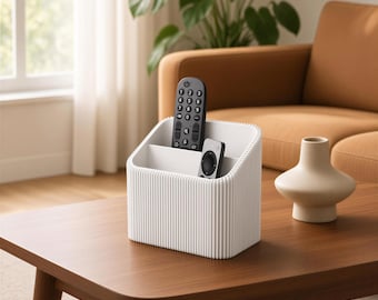 May include: A white, rectangular desk organizer with a ribbed exterior texture and three internal compartments. The organizer holds a black television remote control and a smaller, secondary remote control within its divided sections.
