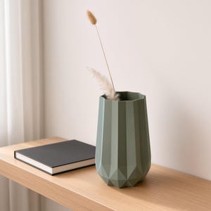Peut inclure: Un vase vert sauge avec une surface extérieure géométrique à facettes. Le vase présente un sommet large et ouvert ainsi qu'une base effilée. À l'intérieur du vase, deux tiges botaniques séchées, duveteuses et claires, s'élèvent de l'ouverture.