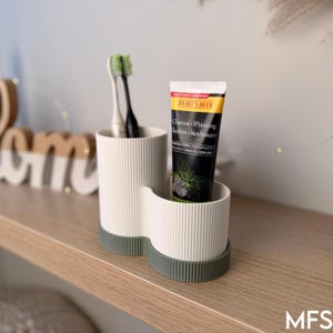 A white and green toothbrush holder with a tube of toothpaste in it, placed on a wooden shelf.
