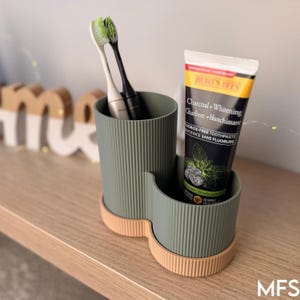 A toothbrush and a tube of Burt's Bees charcoal and whitening toothpaste are placed in a green and beige toothbrush holder on a wooden surface.