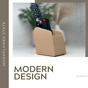 May include: A beige remote control tidy with a black remote inside. The tidy has a minimalist, angled design. The text "MODERN DESIGN" is displayed below the tidy.