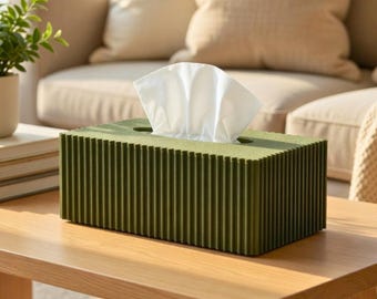 May include: An olive green tissue box with a ribbed design, holding white tissues. The box is on a light wood table. The background features a beige sofa and cushions, creating a neutral, minimalist aesthetic.