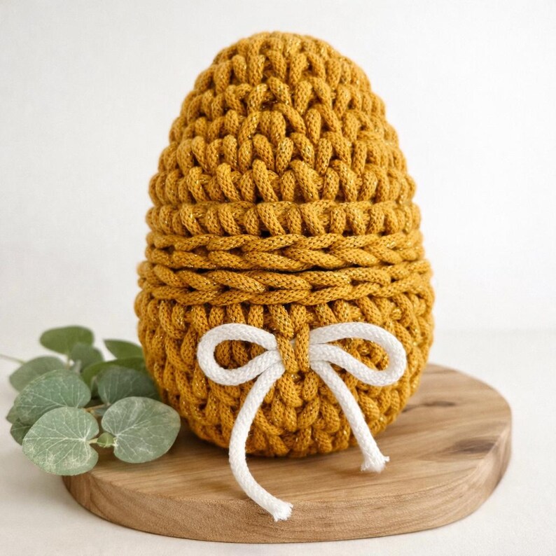 May include: Mustard yellow crocheted egg-shaped basket with a white bow. The basket is made of thick yarn and has a textured appearance. The white bow is tied at the front. The basket is sitting on a wooden surface.
