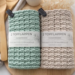 Könnte beinhalten: Zwei handgefertigte Topflappen, einer mintgrün und einer beige, mit einem strukturierten Muster. Jeder Topflappen hat ein Etikett mit der Aufschrift "2 TOPFLAPPEN HANDMADE 100% RECYCELTE BAUMWOLLE HAKELKRAM BY MONIK."