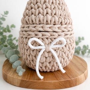 May include: A beige, egg-shaped crochet basket with a white bow. The basket is made of thick, textured yarn and sits on a round wooden board. The top half of the basket is removable. Green foliage is in the background.