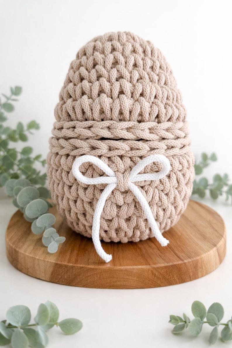 May include: A beige, egg-shaped crochet basket with a white bow. The basket is made of thick, textured yarn and sits on a round wooden board. The top half of the basket is removable. Green foliage is in the background.