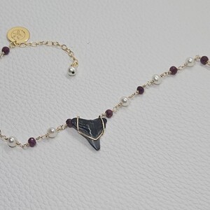 May include: A gold chain bracelet with white pearls and burgundy beads. A shark tooth pendant hangs from the center of the bracelet.