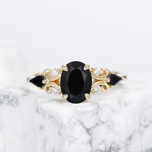 Pode incluir: Um anel de ouro com uma gema preta ovalada cercada por gemas brancas menores. O anel tem um design delicado e ornamentado.