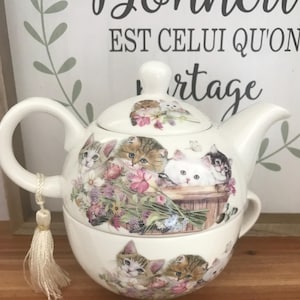 Op de afbeelding: Een wit keramisch theeservies met katten- en bloemmotieven. De theepot heeft een deksel en een handvat met een kwastje. De theekop is onder de theepot gestapeld. De achtergrond heeft Franse tekst.