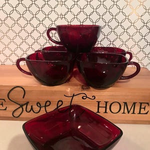 May include: A collection of ruby red glass teacups and a square bowl. The teacups are stacked, with handles visible. The bowl is in the foreground. The background features a wooden sign with the words "Sweet Home" and a patterned wallpaper.