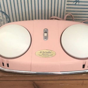 May include: A vintage Dr. Scholl's Electric Foot Massager in a soft pink color. The massager has two large white circular pads, an on/off switch, and a silver trim. The cord and plug are also pink.