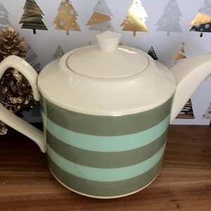 Può includere: Una teiera in ceramica con coperchio e manico color crema e un beccuccio. Il corpo della teiera è decorato con strisce orizzontali verde salvia e azzurro chiaro. La teiera è su una superficie di legno.