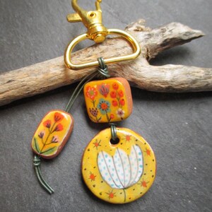 K&ouml;nnte beinhalten: Ein handgefertigter Schl&uuml;sselanh&auml;nger mit einem goldfarbenen Verschluss und drei dekorativen Keramikteilen. Die Teile sind mit floralen Designs in Orange, Gelb und Blau bemalt. Der Schl&uuml;sselanh&auml;nger ist an einem St&uuml;ck Treibholz befestigt.