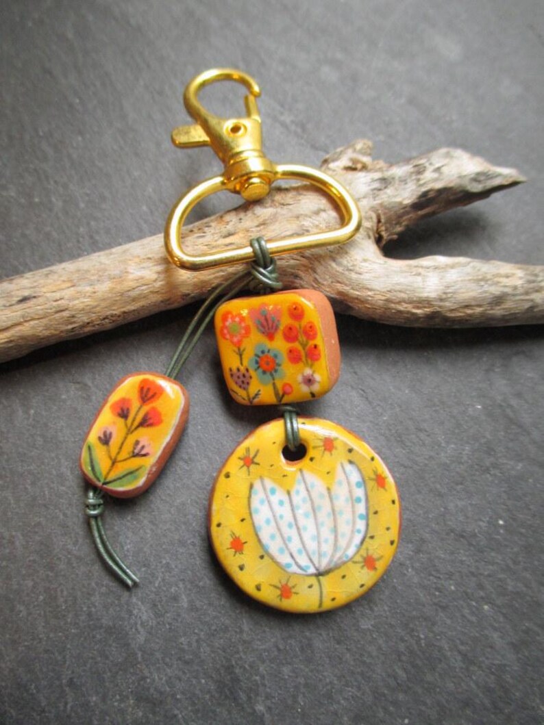 K&ouml;nnte beinhalten: Ein handgefertigter Schl&uuml;sselanh&auml;nger mit einem goldfarbenen Verschluss und drei dekorativen Keramikteilen. Die Teile sind mit floralen Designs in Orange, Gelb und Blau bemalt. Der Schl&uuml;sselanh&auml;nger ist an einem St&uuml;ck Treibholz befestigt.