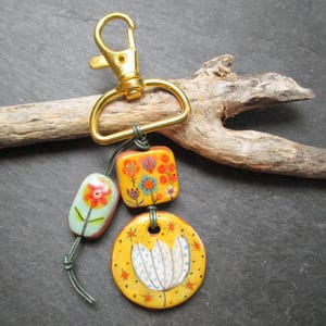 K&ouml;nnte beinhalten: Ein handgefertigter Schl&uuml;sselanh&auml;nger mit einem goldfarbenen Verschluss und drei dekorativen Keramikperlen. Die Perlen sind gelb, orange und blau und weisen florale Designs auf. Der Schl&uuml;sselanh&auml;nger ist an einem St&uuml;ck Treibholz befestigt.