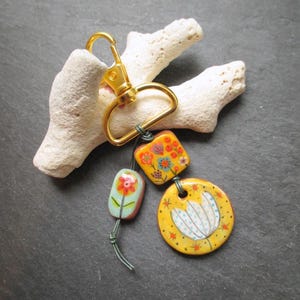 K&ouml;nnte beinhalten: Ein goldfarbener Schl&uuml;sselanh&auml;nger mit drei dekorativen Keramik-Anh&auml;ngern. Die Anh&auml;nger sind mit floralen Designs in Orange, Gelb und Blau bemalt. Der Schl&uuml;sselanh&auml;nger ist an einem wei&szlig;en, strukturierten Treibholzst&uuml;ck befestigt.