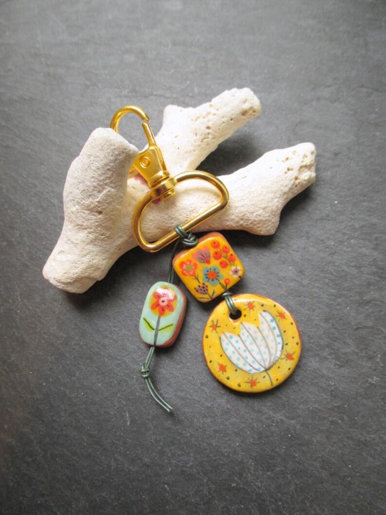 K&ouml;nnte beinhalten: Ein goldfarbener Schl&uuml;sselanh&auml;nger mit drei dekorativen Keramik-Anh&auml;ngern. Die Anh&auml;nger sind mit floralen Designs in Orange, Gelb und Blau bemalt. Der Schl&uuml;sselanh&auml;nger ist an einem wei&szlig;en, strukturierten Treibholzst&uuml;ck befestigt.