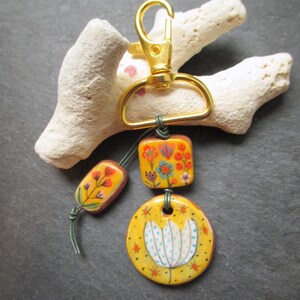 K&ouml;nnte beinhalten: Ein handgefertigter Schl&uuml;sselanh&auml;nger mit einem goldfarbenen Verschluss und drei dekorativen Keramikteilen. Die Teile sind gelb mit floralen Designs in Orange, Blau und Wei&szlig;. Der Schl&uuml;sselanh&auml;nger ist an einem St&uuml;ck wei&szlig;em Korallenriff befestigt.