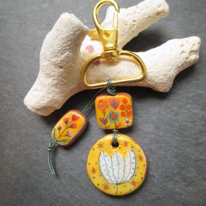 K&ouml;nnte beinhalten: Ein handgefertigter Schl&uuml;sselanh&auml;nger mit einem goldfarbenen Verschluss und drei dekorativen Keramikteilen. Die Teile sind gelb mit floralen Designs in Orange, Blau und Wei&szlig;. Der Schl&uuml;sselanh&auml;nger ist an einem St&uuml;ck wei&szlig;em Korallenriff befestigt.