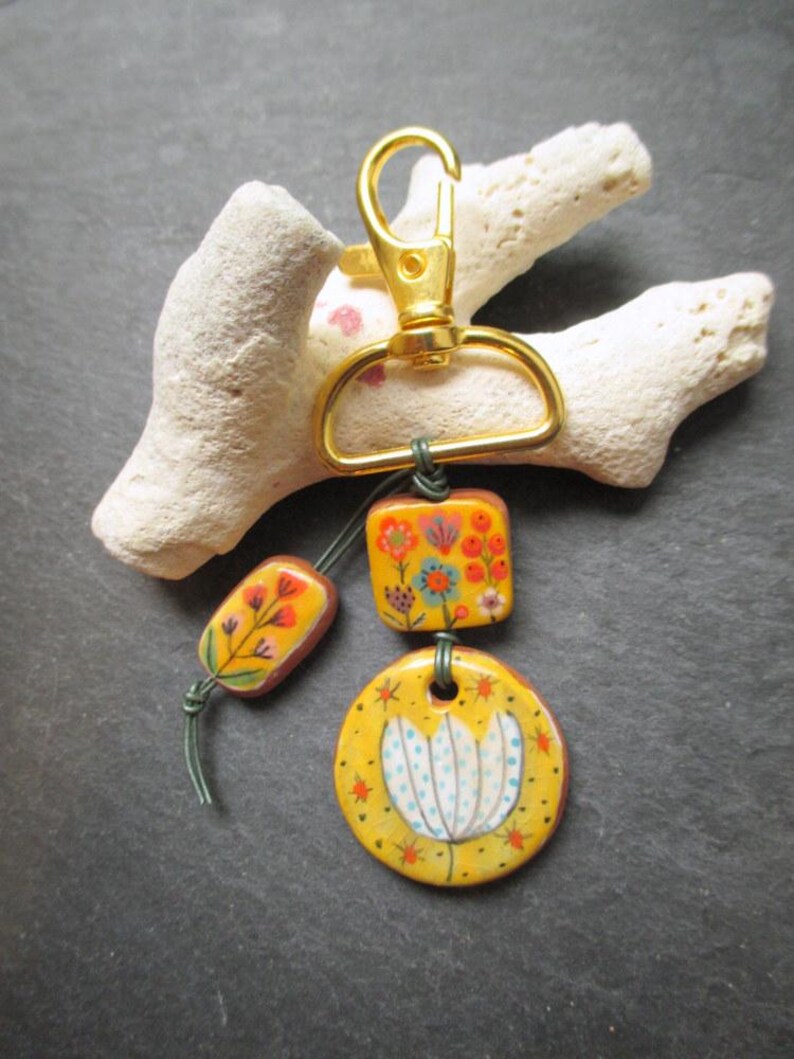 K&ouml;nnte beinhalten: Ein handgefertigter Schl&uuml;sselanh&auml;nger mit einem goldfarbenen Verschluss und drei dekorativen Keramikteilen. Die Teile sind gelb mit floralen Designs in Orange, Blau und Wei&szlig;. Der Schl&uuml;sselanh&auml;nger ist an einem St&uuml;ck wei&szlig;em Korallenriff befestigt.