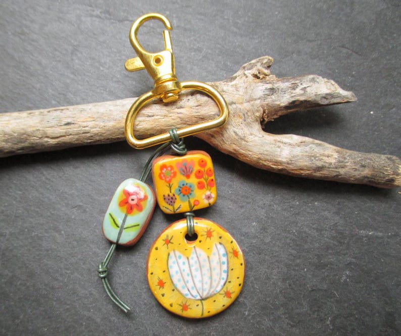 K&ouml;nnte beinhalten: Ein handgefertigter Schl&uuml;sselanh&auml;nger mit einem goldfarbenen Karabinerhaken und drei dekorativen Keramikperlen. Die Perlen sind gelb, orange und hellblau und weisen florale Designs in verschiedenen Farben auf. Der Schl&uuml;sselanh&auml;nger ist an einem St&uuml;ck Treibholz befestigt.