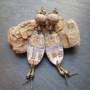 May include: A pair of dangle earrings with oval-shaped pendants featuring a vintage design and gold accents. The pendants have a white background with brown and gold details and text that reads "Fear not" and "On no Ist". The earrings have brown and gold beads hanging from the bottom.