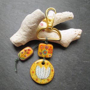 K&ouml;nnte beinhalten: Ein handgefertigter Schl&uuml;sselanh&auml;nger mit einem goldfarbenen Verschluss und drei dekorativen Keramikperlen. Die gelben Perlen sind handbemalt mit floralen Designs in Rot, Orange, Blau und Wei&szlig;. Der Schl&uuml;sselanh&auml;nger liegt auf wei&szlig;em Korallen.