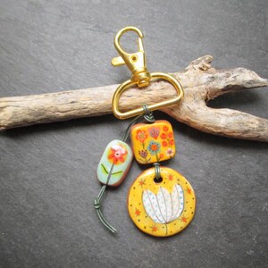 K&ouml;nnte beinhalten: Ein dekorativer Schl&uuml;sselanh&auml;nger mit einem goldfarbenen Verschluss und drei bemalten Keramikanh&auml;ngern. Die Anh&auml;nger zeigen florale Designs in Orange, Gelb und Blau. Der Schl&uuml;sselanh&auml;nger wird auf einem St&uuml;ck Treibholz pr&auml;sentiert.