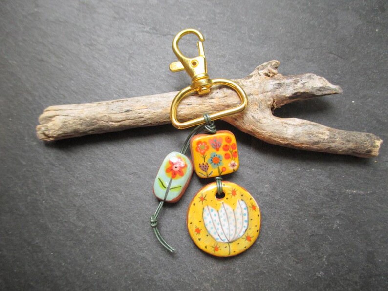 K&ouml;nnte beinhalten: Ein dekorativer Schl&uuml;sselanh&auml;nger mit einem goldfarbenen Verschluss und drei bemalten Keramikanh&auml;ngern. Die Anh&auml;nger zeigen florale Designs in Orange, Gelb und Blau. Der Schl&uuml;sselanh&auml;nger wird auf einem St&uuml;ck Treibholz pr&auml;sentiert.
