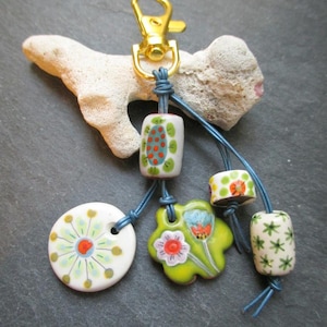May include: A handmade keychain with a gold-colored clasp and decorative ceramic beads. The beads are white, green, red, and blue, featuring floral and geometric designs. The keychain is attached to a piece of coral.