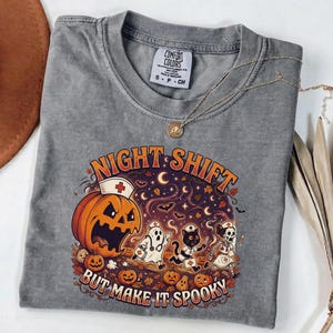 May include: A grey t-shirt featuring a graphic print with the text "NIGHT SHIFT BUT MAKE IT SPOOKY". The design includes a jack-o'-lantern wearing a nurse cap, a ghost, a black cat, and a skeleton, all dressed as medical staff amidst autumn leaves.