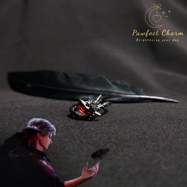 May include: A black ring with a red gemstone and a detailed design of a bird with outstretched wings. The ring is on a black feather and a black background.