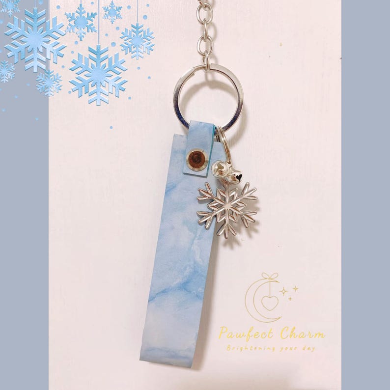 May include: A keychain with a blue marbled leather tag, a silver snowflake charm, and a small bell. The tag is attached to a silver keyring with a metal snap. The background has a light blue and white gradient with snowflake decorations.